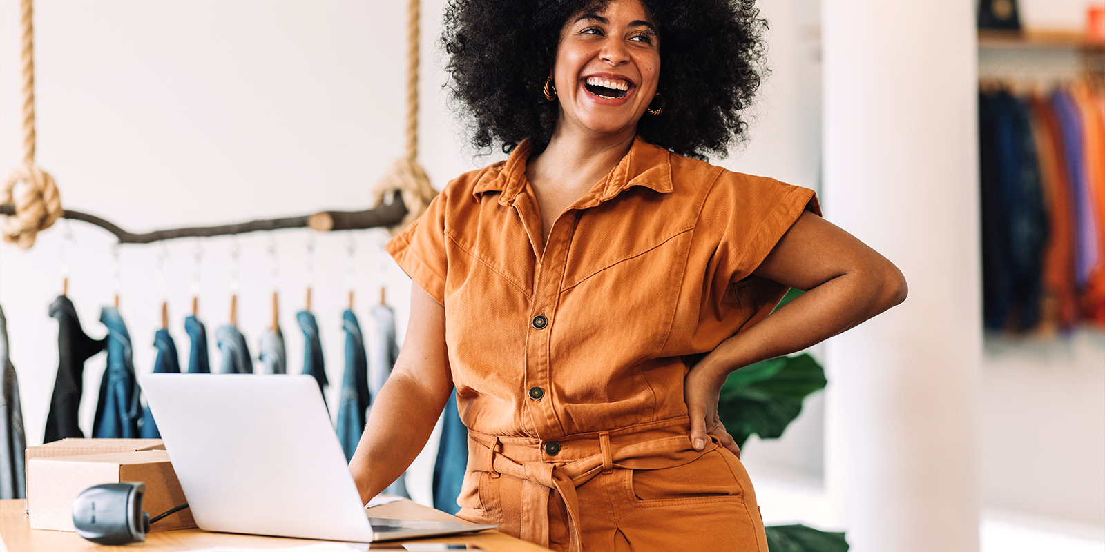 Notre solution Bizgift permet d'accroître votre visibilité en affichant vos cartes sur les sites de notre réseau de distributeurs. image d'une femme souriante dans un commerce avec un ordinateur portable devant elle. En arrière plan, des vêtements sont suspendus.