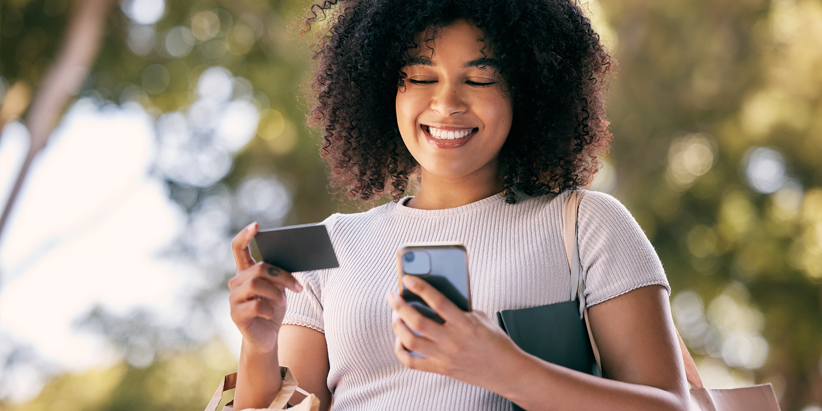 Numen vous accompagne dans la production de cartes cadeaux et cartes prépayées. Image d'une femme, en extérieur, souriant avec un carte cadeau dans une main et une téléphone portable dans l'autre main.