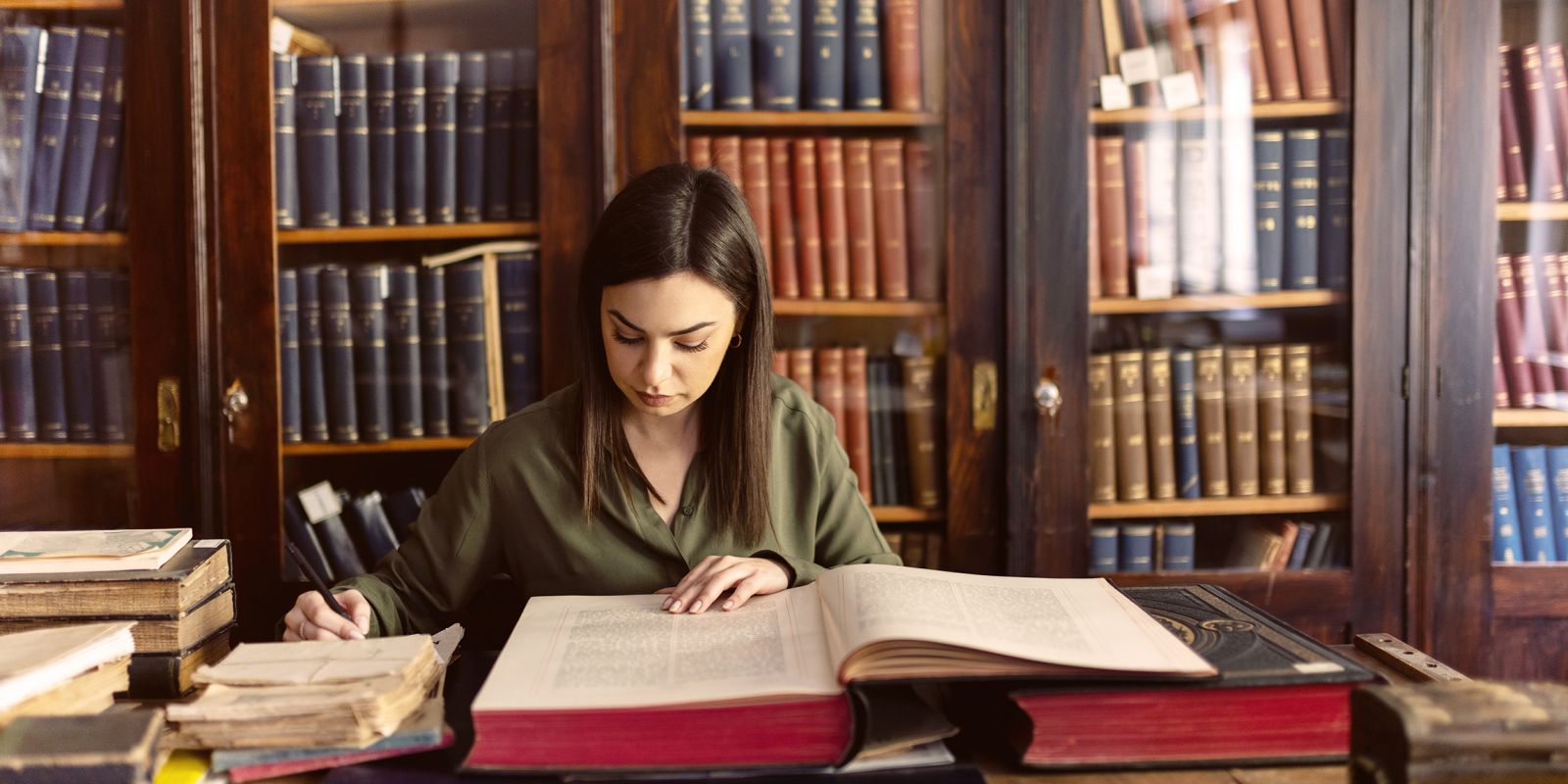 Témoignage client - Chambre des Députés du Luxembourg. Un projet de numérisation d'archives parlementaires. Image d'une femme dans une bibliothèque consultant une grosse archive avec un stylo à la main. Elle est entourée de papiers anciens, de livres précieux et devant une bibliothèque d'ouvrages précieux protégés sous verre.