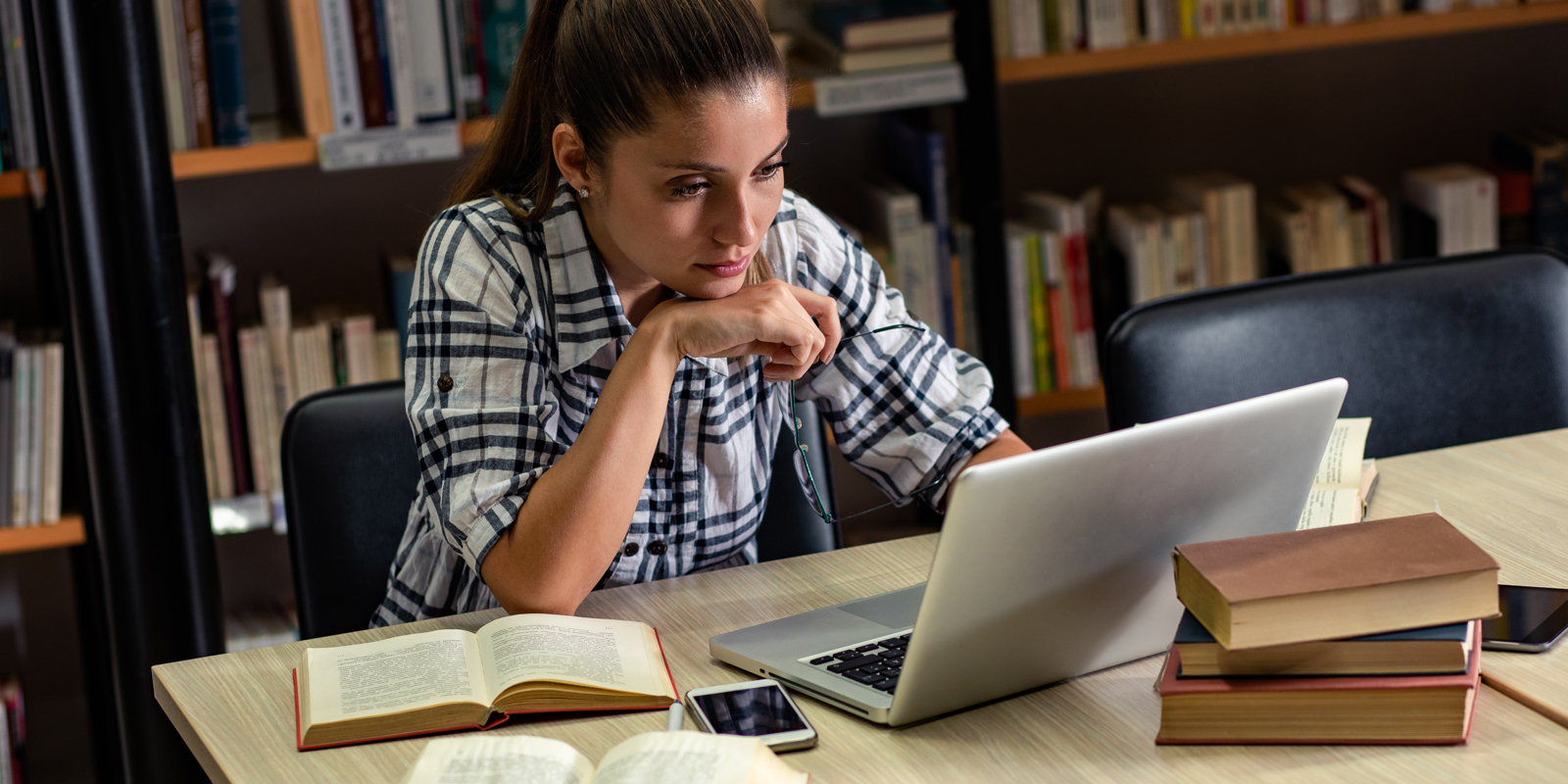 La numérisation patrimoniale et culturelle permet une valorisation des ressources. Image d'une femme dans une bibliothèque avec des ouvrages autour d'elle et un ordinateur portable en face. Image symbolisant une femme travaillant avec des ressources papier aussi accessibles en ligne.